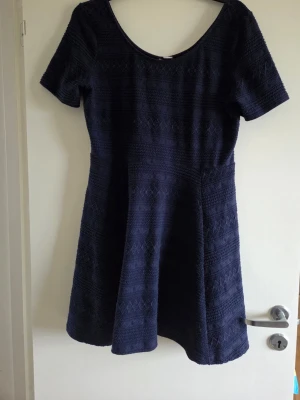 Mörkblå klänning, stl L - Kortärmad mörkblå klänning med rund halsringning och lätt utställd kjol. Klänningen har ett diskret geometriskt mönster i tyget och är tillverkad i ett stretchigt material som ger en bekväm passform. Perfekt för dig som gillar enkel men stilren design.
