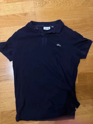 Mörkblå pikétröja från Lacoste - Klassisk mörkblå pikétröja från Lacoste med korta ärmar och den ikoniska gröna krokodilloggan på bröstet. Tröjan har krage och knäppning framtill. Perfekt för en stilren och avslappnad look.