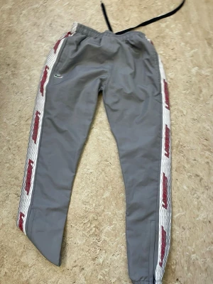 Gråa mjukisbyxor  - Säljer ett par gråa mjukisbyxor från med vita och röda sidopaneler där det står Diadora längs benen. Byxorna har resår i midjan och mudd vid bensluten samt en svart dragsko. Perfekta för chill eller träning.