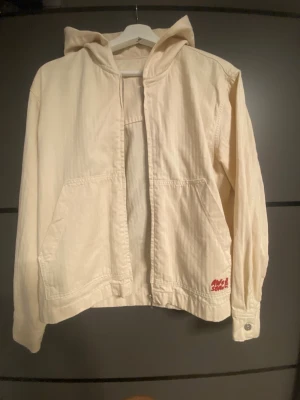 Vit hoodie från Awesome - Säljer en stilren vit hoodie från Awesome med luva och två stora fickor framtill. Hoodien har en avslappnad passform och knappdetaljer vid ärmsluten. Materialet känns mjukt och bekvämt, perfekt för chill dagar. Liten röd logga på ena fickan ger en snygg detalj.