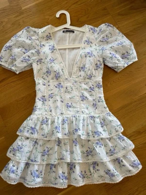 Trendig Zara klänning, Blommig kortklänning med volanger - Supersöt kortklänning med puffärmar, djup v-ringning och flera volanglager. Klänningen är vit med lila och blå blommor, har en feminin passform och är perfekt för sommarens alla tillfällen. Tillverkad i lätt bomullsmaterial som känns luftigt och bekvämt.