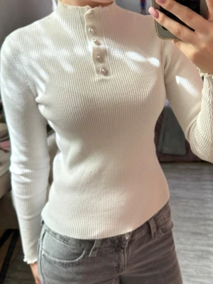 Vit ribbad långärmad topp - Säljer en vit ribbad långärmad topp med hög hals och dekorativa pärlknappar framtill. Tröjan har figurnära passform och lätt vågiga kanter vid ärmslut och hals. Perfekt att styla med jeans eller kjol för en clean look.