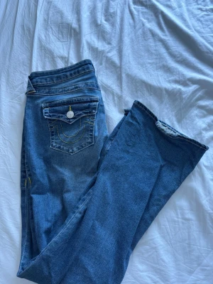 Blå bootcut jeans från True Religion - Säljer ett par blå bootcut jeans från True Religion, modell BECCA Mid Rise Bootcut. Jeansen har snygga kontrastsömmar i gult och klassisk fickdesign med lock och knapp bak. Perfekta för dig som gillar en avslappnad men trendig look.