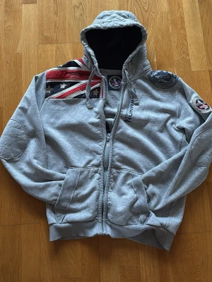 Grå hoodie med patchar och dragkedja - Snygg grå hoodie från Geographical Norway med dragkedja, huva och coola patchar samt broderade detaljer på axel och ärm. Union Jack-flagga på bröstet och quiltad huva. Perfekt för en avslappnad streetstil. Storlek M men sitter som S, därför jag säljer den den passar inte riktigt på mig.