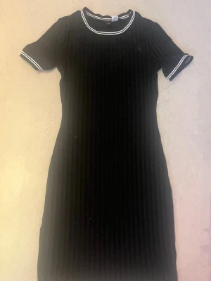 Svart ribbad klänning från H&M Divided - Svart ribbad kortklänning från H&M Divided med vita ränder på krage och ärmslut. Klänningen har korta ärmar och en rund halsringning med sportig känsla. Perfekt för en stilren och enkel look. Knappt andvänd 