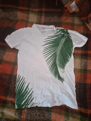 Vit t-shirt med palmblad från Tati - Säljer en vit t-shirt från Tati med grönt palmbladsmotiv på framsidan. Storlek S