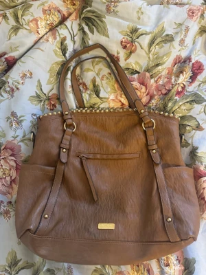 Brun handväska från Jessica Simpson - Snygg brun handväska i fejkläder från Jessica Simpson. Väskan har guldfärgade detaljer, dubbla handtag, dragkedjeficka framtill och dekorativa nitar längs ovankanten. Perfekt för dig som vill ha en rymlig och trendig väska med många fack.