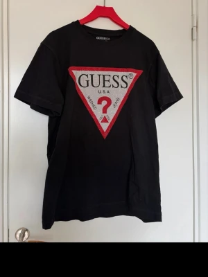 Svart Guess t-shirt med stor logga - Svart t-shirt från Guess med ikonisk stor trekantig logga framtill i vitt och rött. Klassisk passform och korta ärmar. Tillverkad i mjuk bomull som känns skön mot huden. Perfekt för dig som vill ha en statement-tee med streetkänsla.