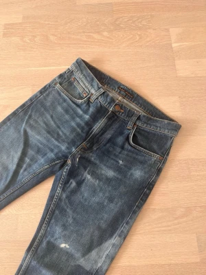 Blå jeans från Nudie Jeans - Säljer ett par klassiska blå jeans från Nudie Jeans med snygg slitning och raka ben. 