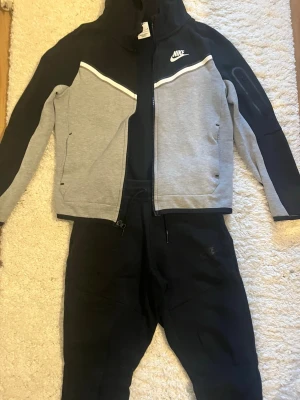 Nike svart och grått loungewear-set - Nike tech set från nike. Tröjan är grå svart och byxorna är hel svarta med dragkedja på sidan av byxorna 