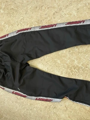 Svarta Lacoste mjukisbyxor med revärer - Svarta mjukisbyxor från Lacoste med vita och röda revärer längs sidorna där det står 'Lacoste'. Byxorna har en avslappnad passform och resår i midjan. Perfekta för en sportig och chill stil.