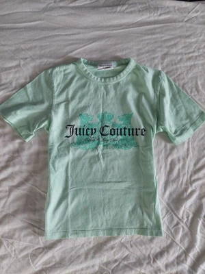 Mintgrön Juicy Couture t-shirt - Snygg mintgrön t-shirt från Juicy Couture med tryck framtill i svart och turkost. Klassisk rund halsringning och korta ärmar. Passar xs/s.