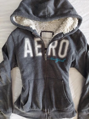 Grå hoodie från Aeropostale - Mysig grå hoodie från Aeropostale med dragkedja och stor huva fodrad med vit fluffig teddy. Stora vita bokstäver 'AERO' och blå text 'New York' på bröstet. Två fickor framtill och ribbade muddar vid ärmar och nederkant. Perfekt för chill dagar.