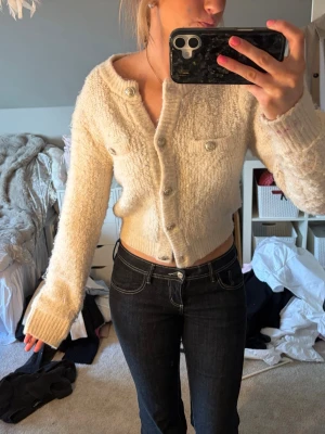 Krämvit kofta från H&M med knappar - Mysig krämvit kofta från H&M med dekorativa silverknappar och två fejkade bröstfickor. Kortare modell med lång ärm och ribbade muddar. Tillverkad i ett fluffigt, stickat material som känns både trendigt och bekvämt.