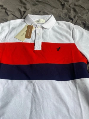 Vit, röd och marinblå pikétröja  - Säljer en stilren pikétröja från Zerone med breda ränder i vitt, rött och marinblått. Tröjan har klassisk krage, knappar framtill och en liten broderad logga på bröstet. Perfekt för dig som gillar sportig och fräsch stil. Pier one, jättefin tröja helt nya. 