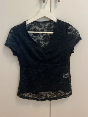 Svart spetsblus med v-ringning - Snygg svart blus i genombruten spets med v-ringad hals och korta ärmar. Toppen har en feminin och elegant look med transparenta detaljer och figurnära passform. Perfekt för dig som vill ha en trendig och stilren topp.