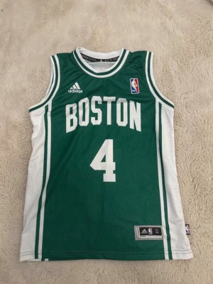 Boston Celtics basketlinne #4 Adidas - Grönt basketlinne från Adidas med vita detaljer och texten 'BOSTON' samt nummer 4 på framsidan. På baksidan står det 'THOMAS' och nummer 4. Officiell NBA-logga och Celtics-märke. Tillverkat i ventilerande meshmaterial, perfekt för basket eller träning.