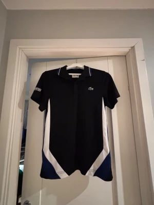 Svart pikétröja från Lacoste - Snygg svart pikétröja från Lacoste med vita och mörkblå detaljer längs sidorna. Klassisk krage, korta ärmar och broderad Lacoste-logga på bröstet. Tillverkad i ett sportigt funktionsmaterial som andas. Perfekt för tennis eller annan träning.
