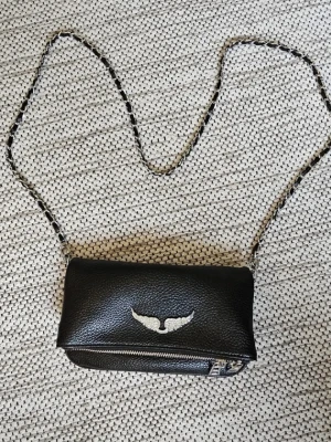 Svart axelväska med kedjerem - Snygg svart axelväska i präg­lat konstläder med silverfärgad kedjerem och ett coolt vitt vingmärke på framsidan. Väskan har dragkedja baktill och praktiskt lock. Perfekt för dig som vill ha en stilren men ändå unik accessoar. EXTRA KEDJA INGÅR, SKRIV OM NI HAR FRÅGOR, PRISET KAN DISKUTERAS. NYPRIS: 4200