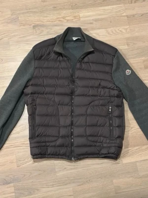 Moncler hybrid pufferjacka grå - Snygg pufferjacka från Moncler i hybridstil med vadderad svart front och grå stickade ärmar och rygg. Jackan har dragkedja, ståkrage och två fickor framtill. Moncler-logga på ena ärmen. Perfekt för kalla vinterdagar. L men passar M