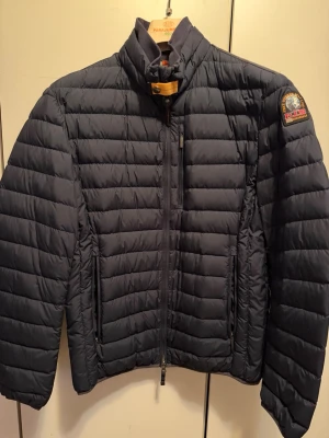 Mörkblå Parajumpers dunjacka - Mörkblå dunjacka från Parajumpers med quiltad design, hög krage och gul detalj vid halsen. Jackan har två sidofickor med dragkedja och en patch med logga på ärmen. Autentisk.