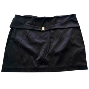 Svart kortkjol med bälte - Stilren svart kortkjol med snyggt bälte och metallspänne framtill. Kjolen har en rak passform och är perfekt för dig som gillar minimalistisk stil. Materialet känns slätt och har en lätt satin finish. Made in Italy.