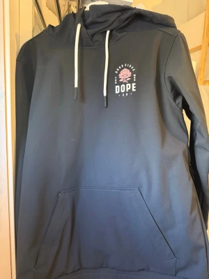 Svart hoodie/slalom vinterjacka från Dope med tryck - Svart hoodie/slalomjacka från Dope med vit dragsko i huvan och ett litet tryck på bröstet med texten 'Good Vibes' och en rosa blomma. Hoodien har en stor magficka och mjukt fleecefoder på insidan. Perfekt för chill dagar och streetwear-stil. Använd 1 gång