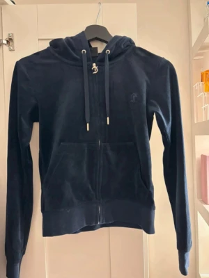 Mörkblå velour hoodie från Juicy Couture - Snygg mörkblå hoodie i mjuk velour från Juicy Couture. Dragkedja framtill, känguruficka och justerbar huva med snören. Perfekt för en chill och trendig look.