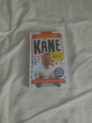 KANE ÄGER - Fotbollsstjärnor - Rolig och inspirerande bok om fotbollsstjärnan Harry Kane, skriven av Simon Mugford och illustrerad av Dan Green. Boken har färgglad framsida med tecknad stil och insidan innehåller både text och svartvita illustrationer. Perfekt för unga fotbollsälskare.
