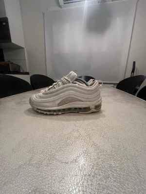 Nike Air Max 97 vita sneakers - Nike Air Max 97 sneakers i vitt med ikoniska vågformade paneler och synlig Air-enhet i sulan. Skorna har reflekterande detaljer, snörning och en sportig siluett. Tillverkade i textil och syntetmaterial för en clean och modern look.