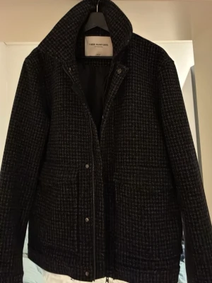 Mörkgrå ulljacka från Three Glory Days - Snygg mörkgrå jacka från Three Glory Days i ullblandning med diskret rutmönster. Klassisk krage, knäppning framtill och två stora fickor. Perfekt för kyliga vår o höst dagar och ger en stilren look. 