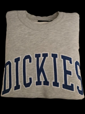 Grå sweatshirt från Dickies - Säljer en grå sweatshirt från Dickies i storlek medium. Tröjan har ett stort blått Dickies-tryck broderat över bröstet och är gjord i mjukt sweatshirtmaterial med ribbade muddar vid hals, ärmslut och nederkant. Perfekt för en avslappnad och stilren look.