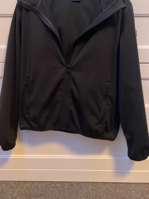 Svart vindjacka med dragkedja - Stilren svart vindjacka med hel dragkedja framtill och två fickor med dragkedja. Jackan har en enkel design och resår vid ärmslut och nederkant. Perfekt för blåsiga dagar och lätt att matcha med olika outfits.