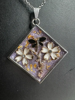 Lila halsband med blommor och trollslända - Unikt halsband med fyrkantigt hänge i silverfärg, dekorerat med två vita blommor, en svart trollslända och gnistrande stenar. Bakgrunden är lila med inslag av guldfärgade detaljer. Perfekt för dig som gillar naturinspirerade smycken.