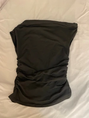 Svart axelbandslös tröja - Säljer en svart tröja med rynkade sidor. Tröjan har en enkel och stilren design som sitter tight och markerar figuren. Perfekt för dig som gillar minimalistisk stil och vill ha något enkelt men snyggt. Vet inte vart jag köpte den det står ej i tröjan.
