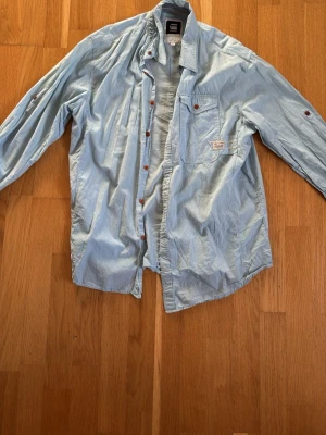 Ljusblå skjorta från G-Star RAW - Säljer en ljusblå skjorta från G-Star RAW med bröstficka och knappstängning. Skjortan har långa ärmar som kan rullas upp och fästas med knapp. Perfekt för en avslappnad och cool stil. Storlek XL men passar perfekt på L.