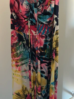 Färgglad långklänning med blommönster - Säljer en klänning med färgstarkt blommönster i rosa, gul, blå och svart. Klänningen har omlottdetalj vid bysten och knytband framtill. Perfekt för dig som vill sticka ut med en unik och somrig look. Jag vet inte storlek men passar kanske M/XL