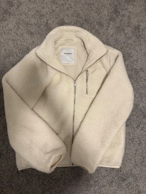 Fleecejacka Jack & Jones - Säljer en fluffig och varm vit fleecejacka från Jack & Jones. Jackan har hel dragkedja framtill, hög krage och en bröstficka med dragkedja. Tillverkad i mjukt teddy-material som ger en riktigt cozy vibe. Enkel att styla till olika outfits.