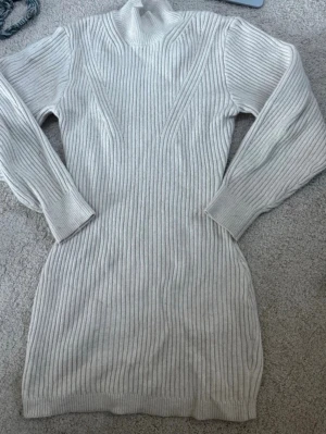 Vit ribbad stickad klänning - Snygg vit ribbad stickad klänning med lång ärm och hög hals. Klänningen har en kroppsnära passform och är perfekt för kyligare dagar. Den ribbade strukturen ger en modern och stilren look.