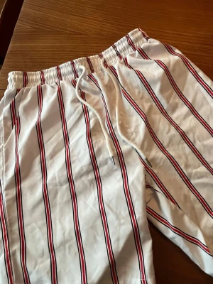 Vita randiga shorts med snörning - Snygga vita shorts med röda och svarta ränder. Shortsen har elastisk midja och snörning framtill för skön passform. Perfekta för sommardagar och avslappnade tillfällen.