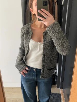 Kofta från Maje - Säljer en gråmelerad kofta från Maje med två svarta fickor framtill och rundad halsringning. Koftan är stickad och har långa ärmar, perfekt att slänga över en topp för en chill look. Materialet är mjukt och bekvämt.