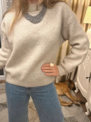 Beige stickad tröja med rund hals - Mysig beige stickad tröja med rund hals och ribbade muddar vid ärmslut och nederkant. Tröjan har en avslappnad passform och är perfekt att matcha med jeans. Materialet ser mjukt och skönt ut, perfekt för kyligare dagar.