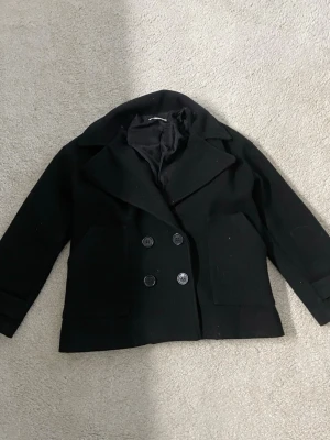 Svart kappa  med dubbelknäppning -  svart kappa med breda slag och dubbelknäppning framtill. Jackan har två stora framfickor och är helfodrad för extra komfort. Klassisk och tidlös modell som passar perfekt till höst och vinter. Materialet känns tjockt och värmande.