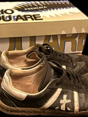 Premiata svarta och vita sneakers - Svarta sneakers från Premiata med vita detaljer och logga på sidan. Skorna har lågt skaft, snörning och en beige insida. Tillverkade i läder med slitstark sula. Perfekta för dig som gillar en klassisk men cool stil.
