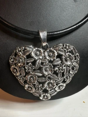 Halsband med hjärthänge i silverfärg - Unikt halsband med ett stort hjärtformat hänge i silverfärgad metall. Hänget har ett detaljerat blommönster med små blommor och blad, vilket ger en vintage och romantisk känsla. Remen är av svatt läder. Perfekt för dig som gillar statement-smycken.
