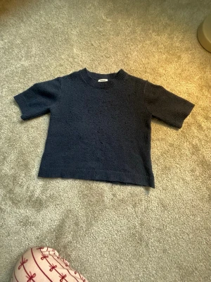 Mörkblå stickad tröja från Kappahl - Säljer en mörkblå stickad t-shirt från Kappahl med korta ärmar och rund halsringning. Tröjan har ribbstickade detaljer vid halsen och nederkanten. Perfekt för dig som gillar en enkel och stilren look.