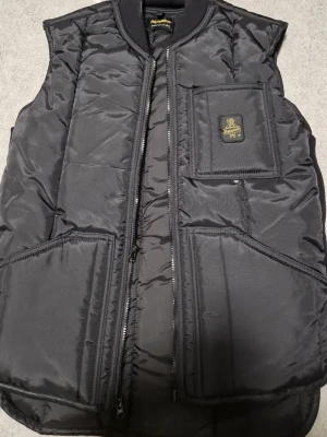 RefrigiWear - Snygg svart dunväst från RefrigiWear med quiltad design och dragkedja framtill. Västen har en bröstficka med patch och två stora fickor framtill. Perfekt för lager på lager och streetwear-stil. Helt ny 