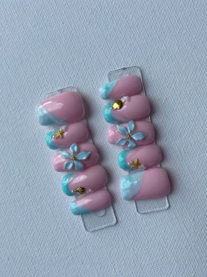 Press-on naglar rosa med blå blommor - Snygga press-on naglar i ljusrosa med blå och vita detaljer. Dekorerade med 3D-blommor, guldfärgade stjärnor och pärlor. Naglarna har en oval form och en glansig finish. Perfekt för dig som vill ha en unik och kreativ nageldesign.