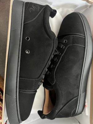 Svarta sneakers från Christian Louboutin - Snygga helsvarta sneakers från Christian Louboutin med klassisk röd sula och signaturdetaljer. Skorna har ovandel i mocka, svarta skosnören och diskreta sömmar. Perfekta för dig som vill ha en clean och exklusiv look.