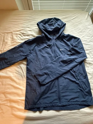 Nike miller - Snygg mörkblå vindjacka från Nike med huva och dragkedja framtill. Jackan har två sidofickor och en diskret Nike-logga på bröstet. Perfekt för blåsiga och blöta dagar och enkel att matcha med olika outfits.